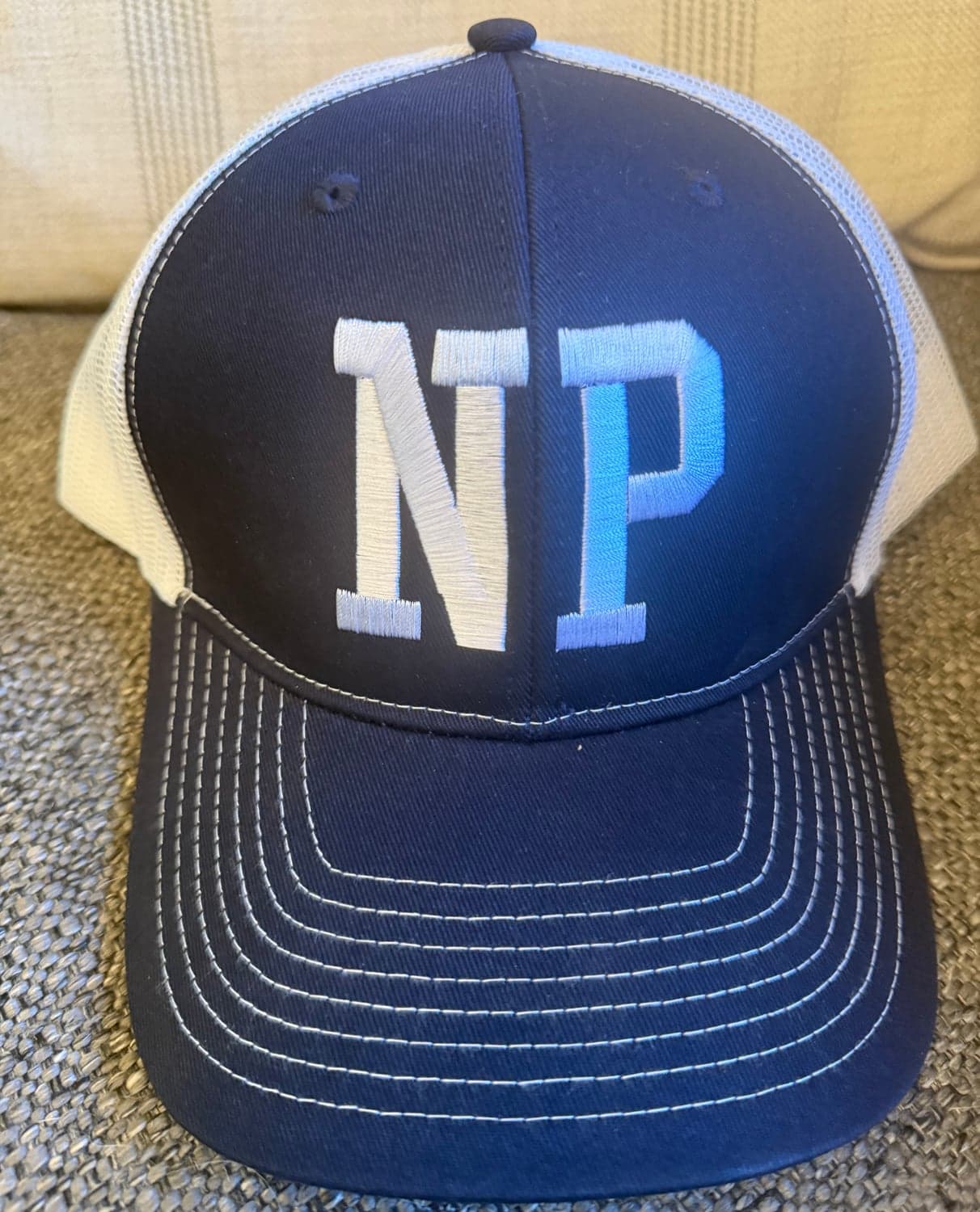 A navy trucker hat with NP embroidered in white on the front, a white mesh back and a navy brim with rows of white stitching.