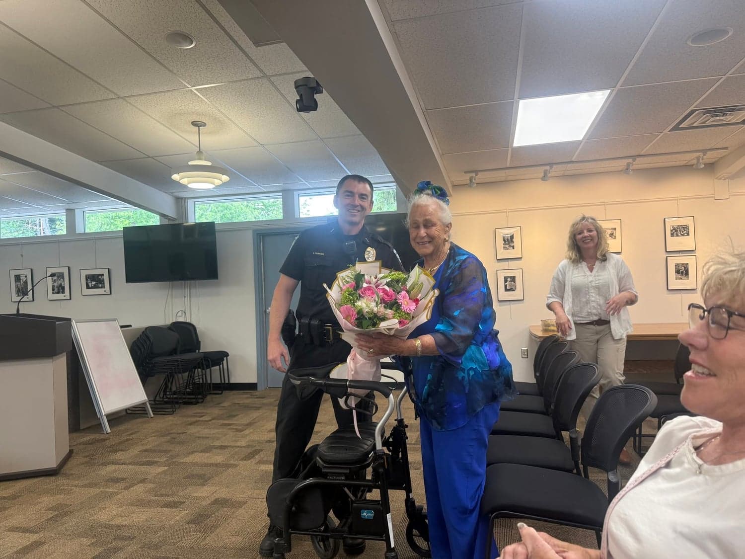 A smiling Normandy Park police officer stands beside a resident holding a bouquet of pink flowers in a meeting room.