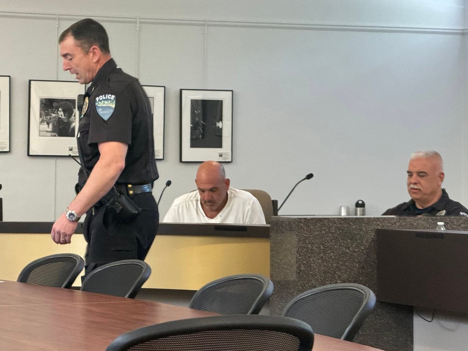 A police officer walks past a meeting table where two men sit at microphones.