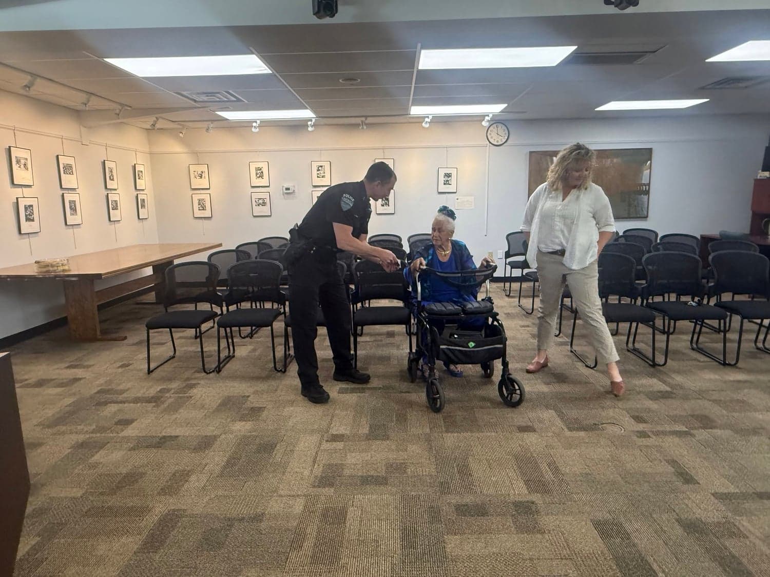 A police officer bends to speak with a seated resident using a walker in a meeting room, as a woman walks over.