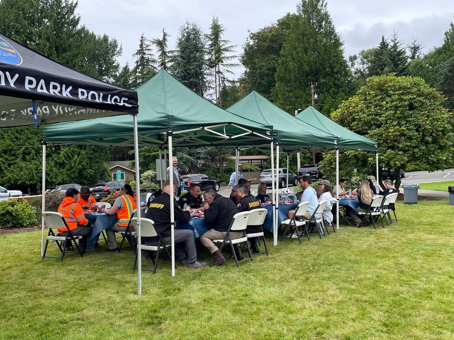 Officers and staff eat lunch at tables under green canopy tents on a lawn, beside a Normandy Park Police canopy.
