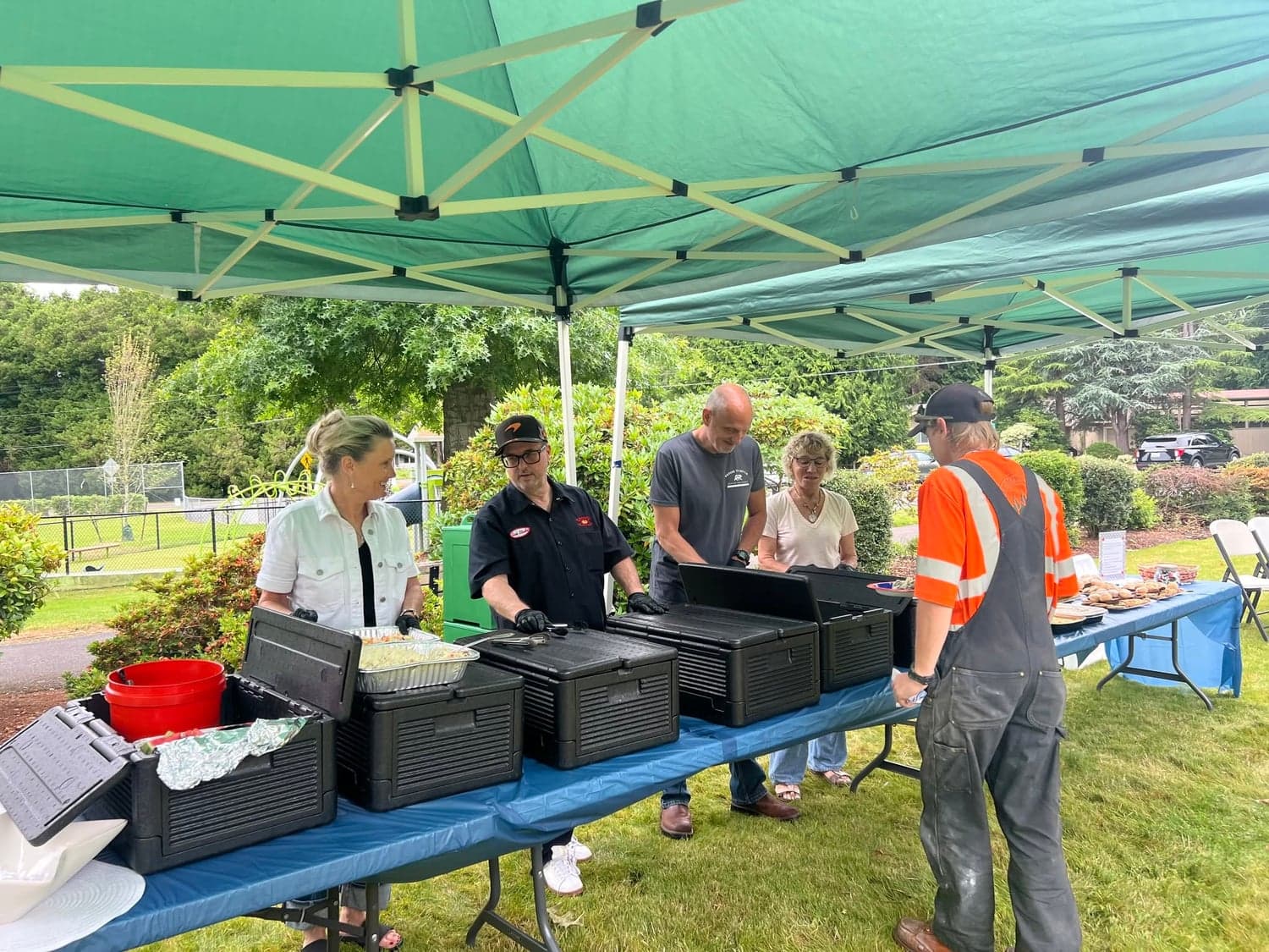 Volunteers in gloves serve food from insulated containers on a buffet table under a green canopy.