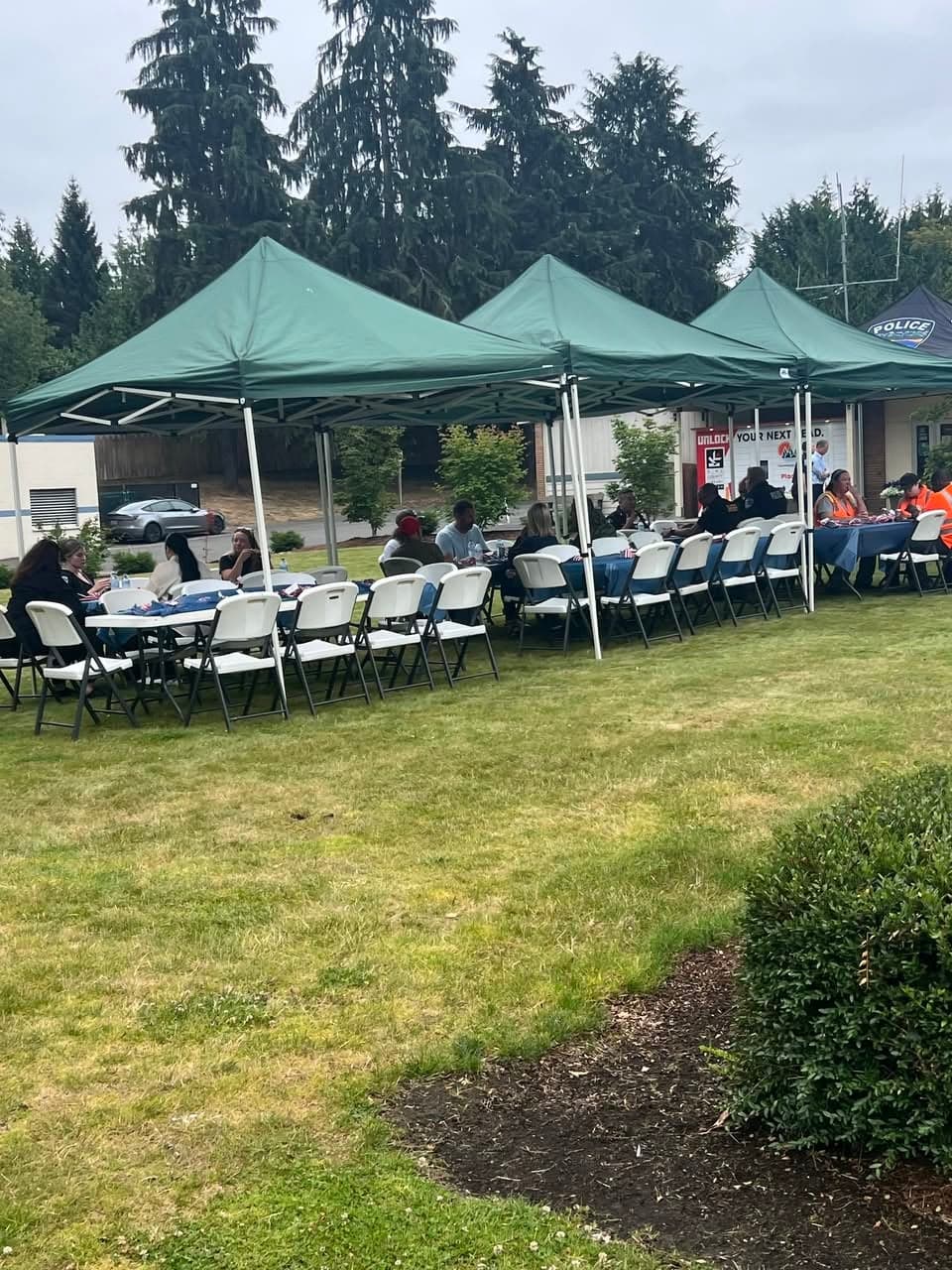 A row of green canopy tents over tables where guests sit and eat, seen across a lawn.