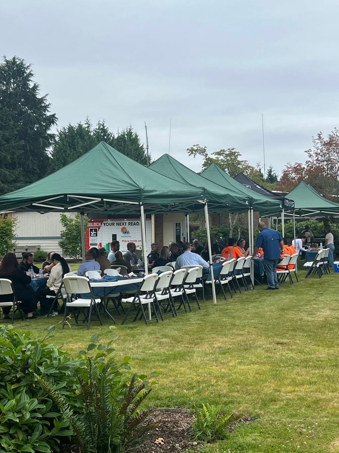 Guests seated at long tables under a row of green canopy tents on a lawn.